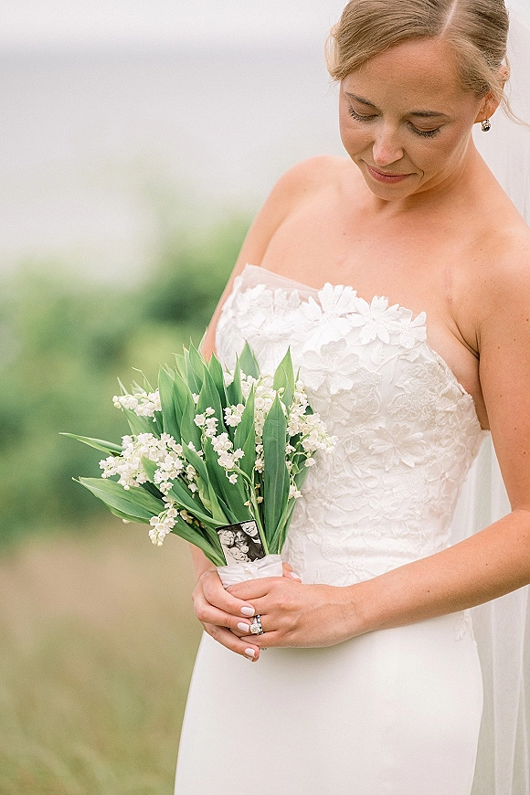 Bridal portrait of a bride holding bouquet of lily of the valley, looking down in a strapless lace dress with veil against blurred greenery