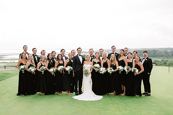 Wedding party photo with bride and groom flanked by bridesmaids in black dresses and tuxedoed groomsmen, oceanview golf lawn under clouds