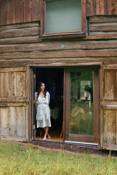 Bridal portrait of a bride in satin robe with lace trim, standing barefoot by a sliding glass door at a rustic wooden cabin exterior
