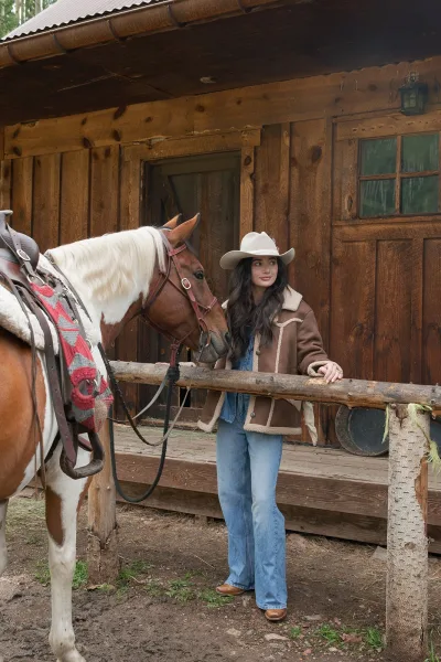 Cowgirl portrait in a white cowboy hat and shearling jacket beside a saddled horse by a rustic wood cabin porch and fence outdoors
