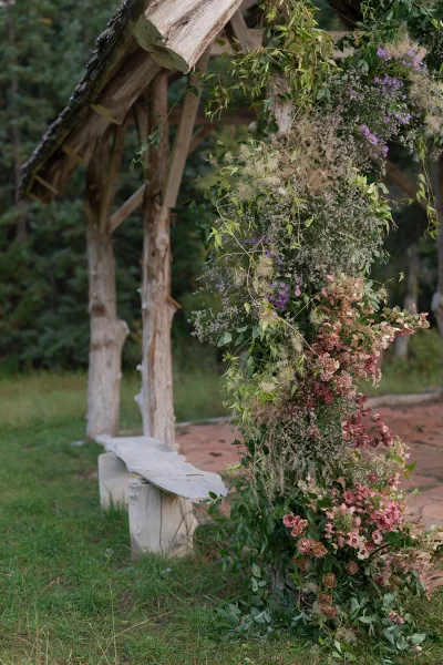 Wedding ceremony arbor with asymmetrical ceremony arch flowers, lush greenery and pink-purple blooms on a grass lawn by trees and brick walkway