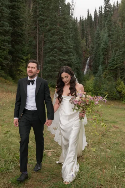 Couple portrait of bride and groom walking hand in hand, bride in long veil with bouquet, beside a waterfall in evergreen meadow forest