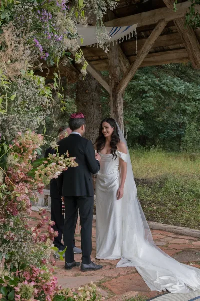 Ceremony moment at a jewish wedding ceremony with bride and groom under a wooden chuppah, long veil and tux, hanging florals in trees
