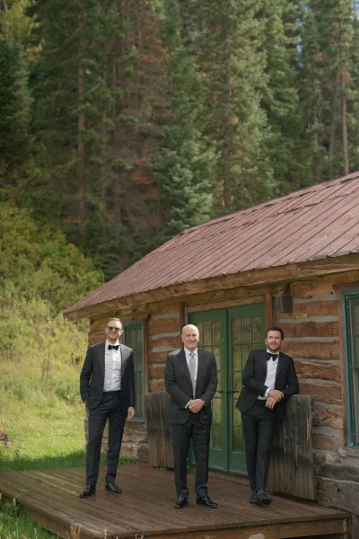 Groomsmen portrait of three men in black tuxedos with bow ties and sunglasses, posing on a log cabin deck with forest behind