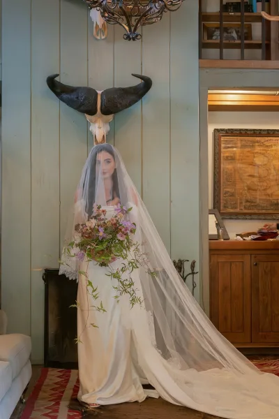Bridal portrait of a cathedral veil bride holding a purple bouquet with greenery, standing by a wood-paneled wall with bull skull decor