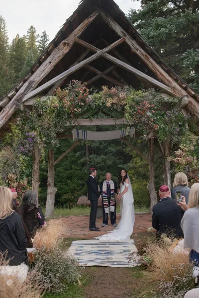 Wedding ceremony with bride and groom at a rustic wooden ceremony arch with floral garland, guests seated on a forest stone patio