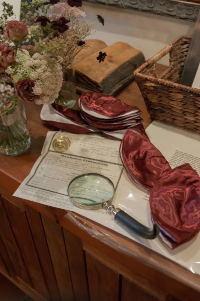 Wedding flat lay of marriage certificate and documents with magnifying glass, burgundy satin kippah and tie on a wood tabletop with vase flowers