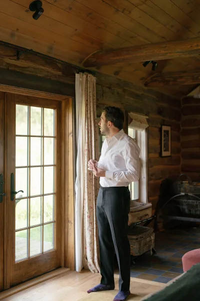 Groom getting ready, buttoning cuffs in a white dress shirt and black pants, standing by a window in a rustic wood cabin interior