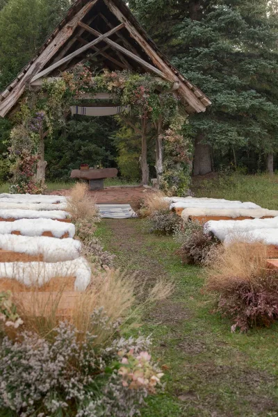 Ceremony setup with a wood arbor and asymmetrical floral arch, striped aisle runner, wooden benches with sheepskin, in a forest clearing
