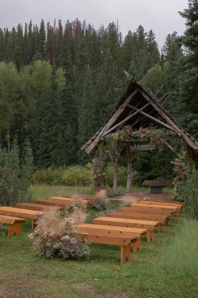 Ceremony setup for an outdoor wedding ceremony with wooden benches facing a rustic altar, framed by greenery garlands in a pine forest meadow