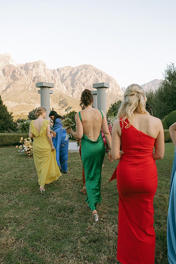 Bridesmaids walking in colorful dresses hold simple bouquet greenery during a garden ceremony entrance with a mountain backdrop
