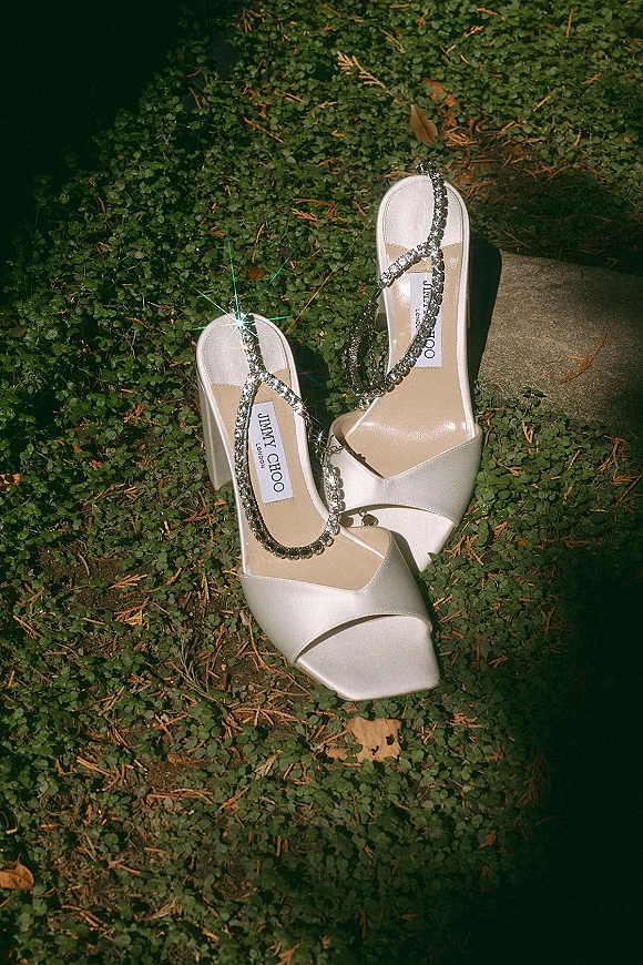 Wedding shoes with rhinestone ankle straps, white satin open-toe block heels resting on green ground cover by a stone step