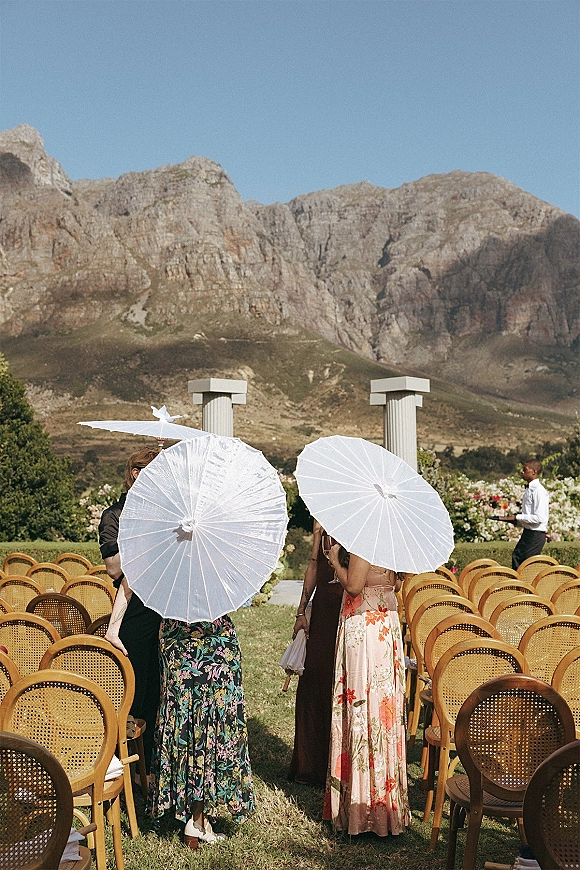 Ceremony seating with outdoor ceremony chairs and cane back wedding chairs lining an aisle runner, guests holding white parasols against mountains
