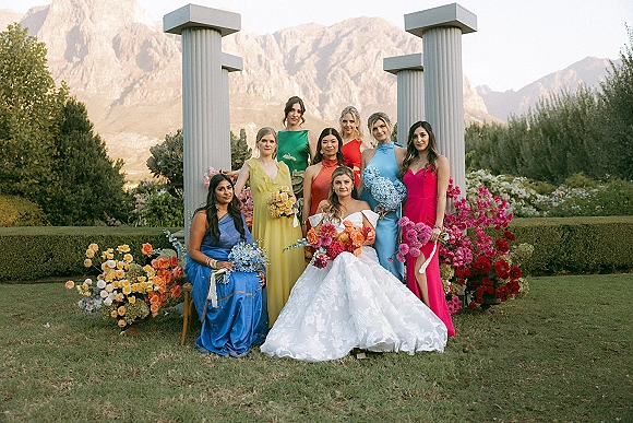 Bridesmaid group portrait with bride with bridesmaids in colorful dresses holding bouquets on a lawn with garden columns and mountains behind