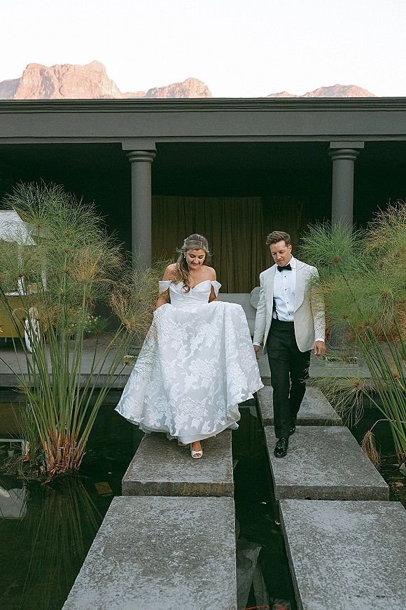 Couple portrait of bride and groom walking hand in hand on stone stepping stones by a reflecting pool with mountains behind