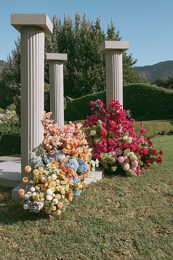 Wedding ceremony backdrop with modern wedding columns topped in rose and hydrangea florals on a lawn with mountains and blue sky behind
