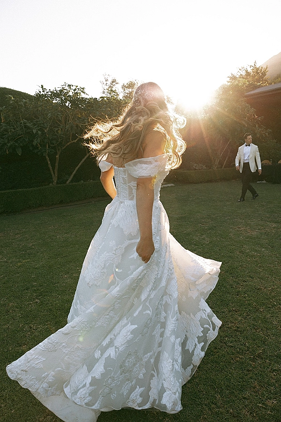 Bridal portrait of a bride twirling in an off shoulder lace wedding dress, groom in white tuxedo behind on a sunset garden lawn