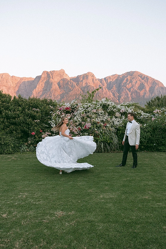 Couple portrait of bride twirling dress as groom watches, her strapless full skirt sweeping across a lawn with mountains behind