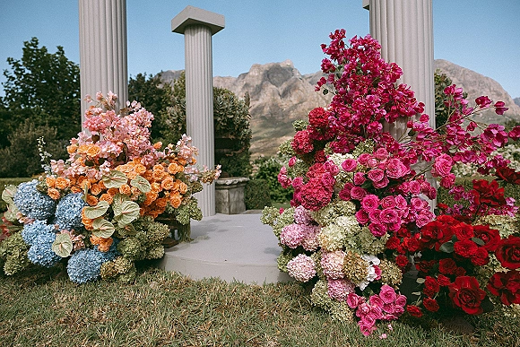 Wedding ceremony floral decor with ceremony altar flowers on white columns, bright roses, hydrangeas and anthurium against a mountain view lawn