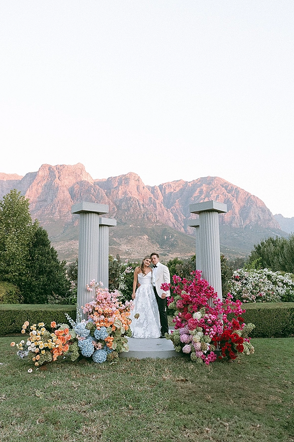 Couple portrait of bride in strapless lace gown and groom in white jacket with bow tie, framed by floral columns and mountain garden backdrop