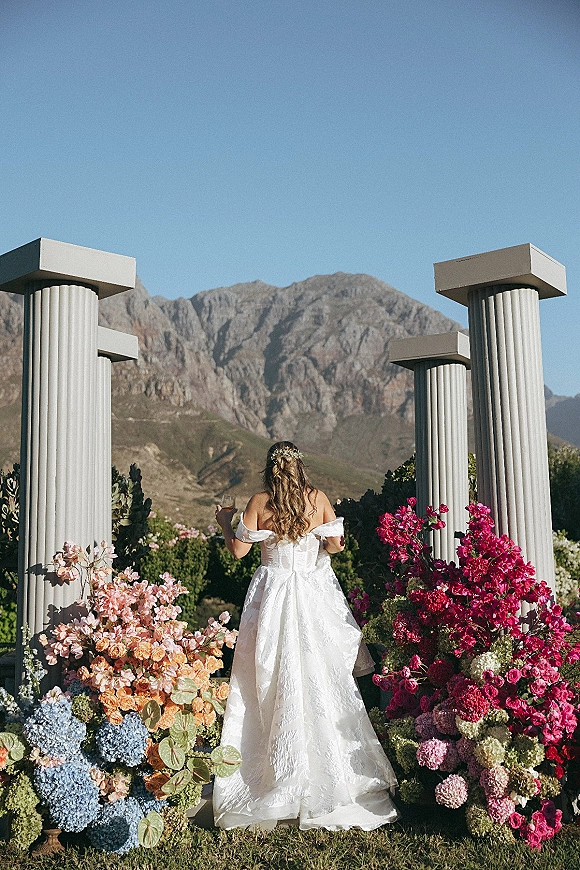 Bridal portrait of a bride from behind holding champagne in an off shoulder wedding dress, facing floral columns with mountains beyond