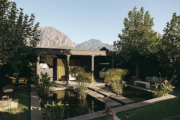 Outdoor lounge setup with pergola curtains, white sofa and cushioned chairs beside a reflecting pool, with mountains beyond the lawn