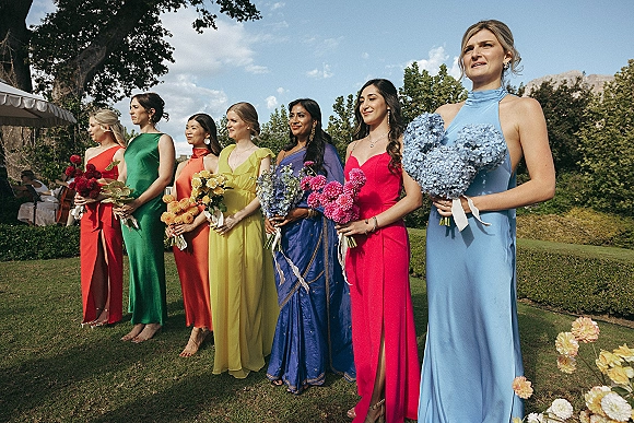 Bridesmaid lineup in colorful dresses holding bouquets with ribbon wraps on a garden lawn under blue sky and a canopy tent