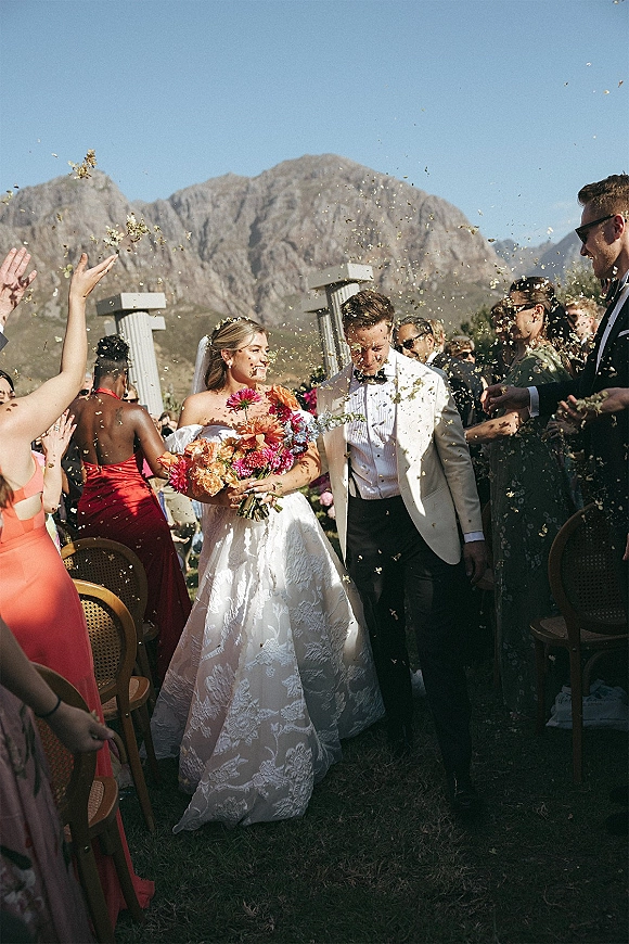 Wedding recessional as bride and groom walk up the aisle through confetti, bride holding bouquet under a blue mountain sky