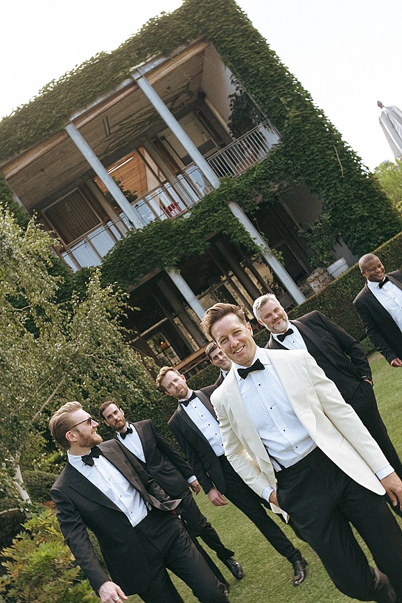 Groomsmen portrait with black tie groomsmen, groom in a white dinner jacket walking on a garden lawn by an ivy-covered building