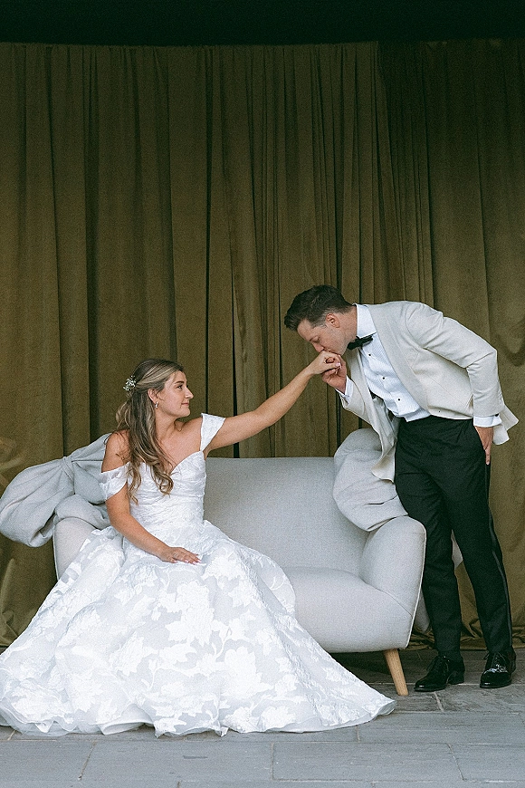 Couple portrait of groom kissing bride’s hand as she sits on an upholstered loveseat, off-the-shoulder gown against draped curtains