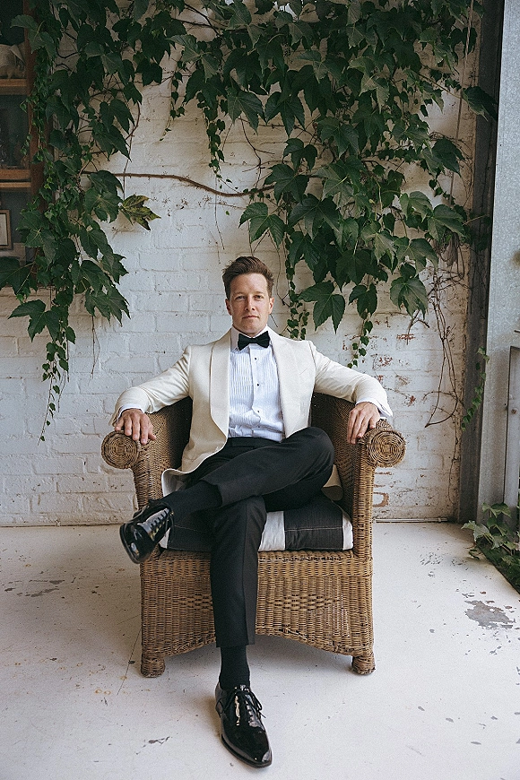 Groom portrait of a man in a white dinner jacket with black bow tie, seated in a wicker armchair against an ivy-covered brick wall