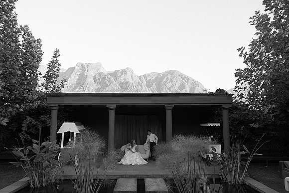 Couple portrait in a black and white wedding photo, bride in strapless gown seated on outdoor lounge sofa as groom stands by pavilion columns with mountains behind