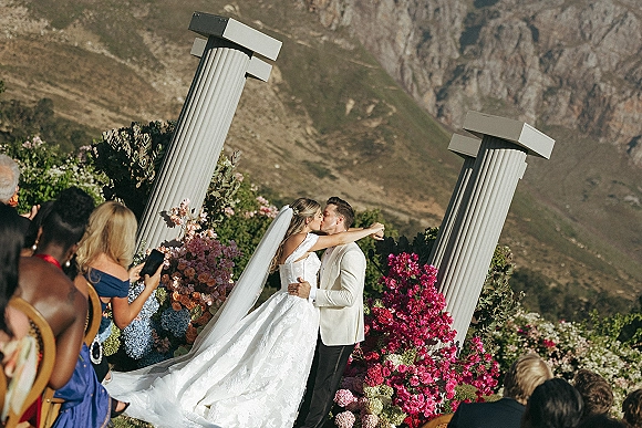 Wedding kiss beneath ceremony columns and hydrangea rose floral arch, bride’s long veil and groom in suit with mountains behind