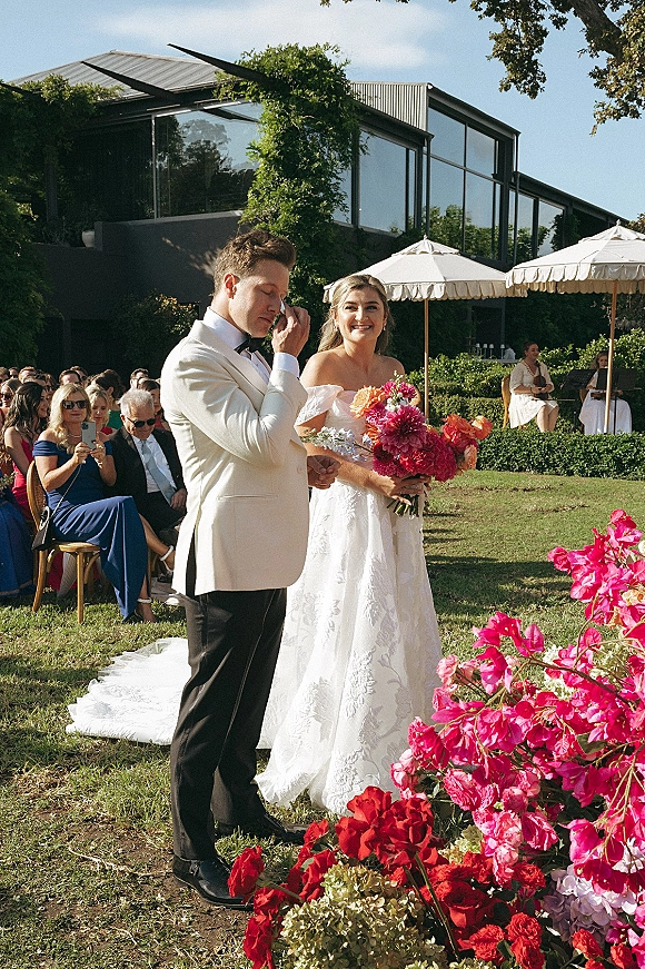 Ceremony moment as groom cries at altar beside smiling bride with bright pink bouquet, under veil on lawn with guests and glass building behind