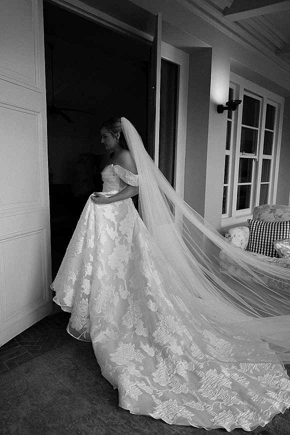 Bridal portrait in black and white of a bride standing in a doorway, wearing an off-the-shoulder jacquard gown with long veil and train on a tiled porch