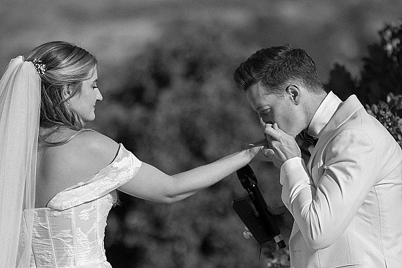 Ceremony moment of groom kissing the bride’s hand during wedding vows, bride in long veil and off-shoulder dress beside trees and sky
