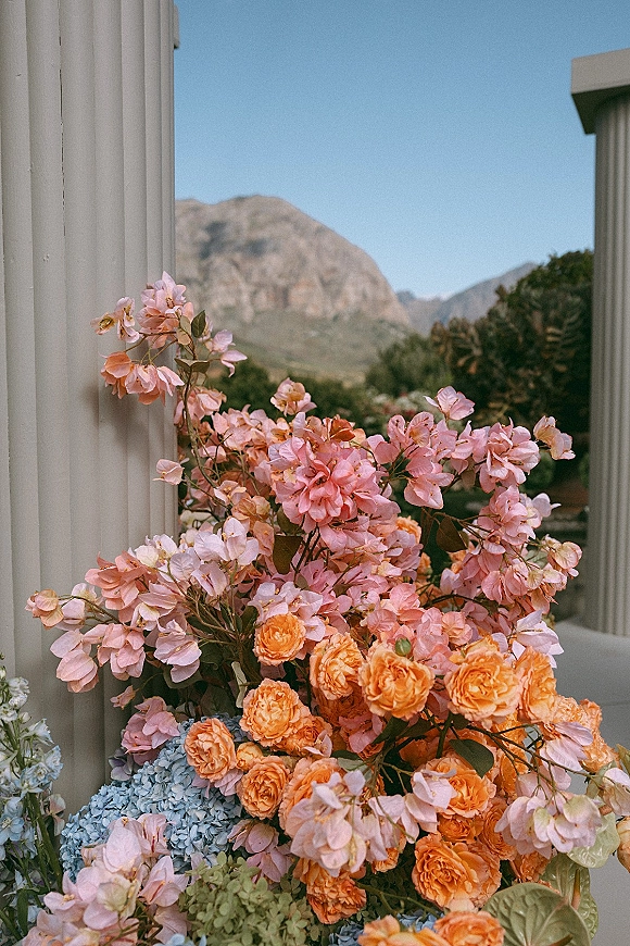 Wedding floral arrangement with pink bougainvillea and orange roses atop white columns, set against mountains, trees, and blue sky