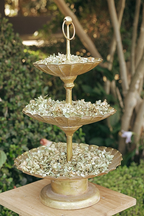 Wedding flower stand on a gold tiered stand filled with dried petals, set on a wooden table amid garden greenery and tree trunks