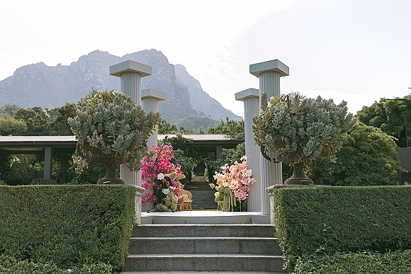 Ceremony backdrop with white and pink floral arrangements on columns and a flower arch, set on garden steps with mountain view beyond