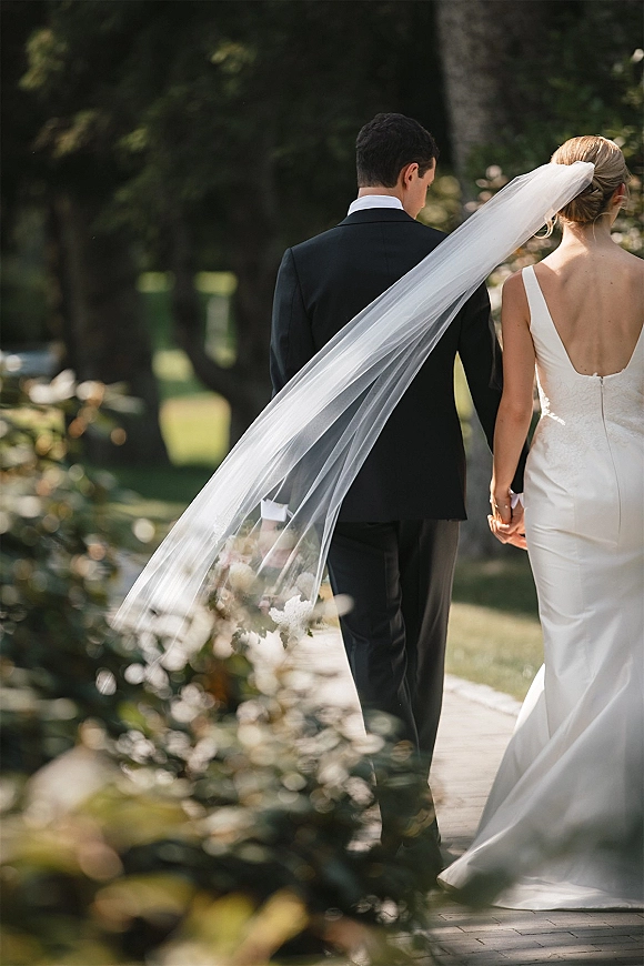 Couple portrait of bride and groom walking away holding hands, long wedding veil trailing on a tree-lined garden pathway