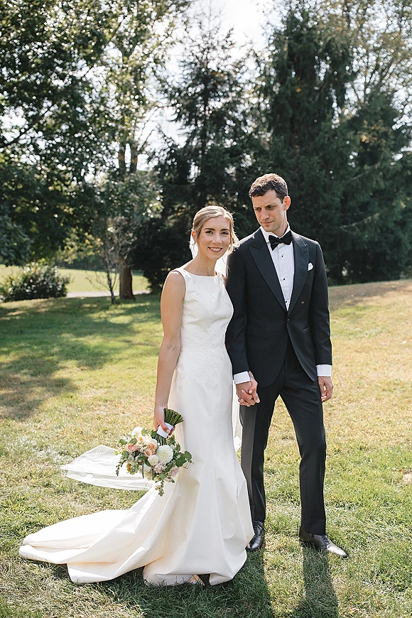 Couple portrait of bride and groom holding hands on a grassy lawn, her veil and bouquet with greenery beside his black tuxedo