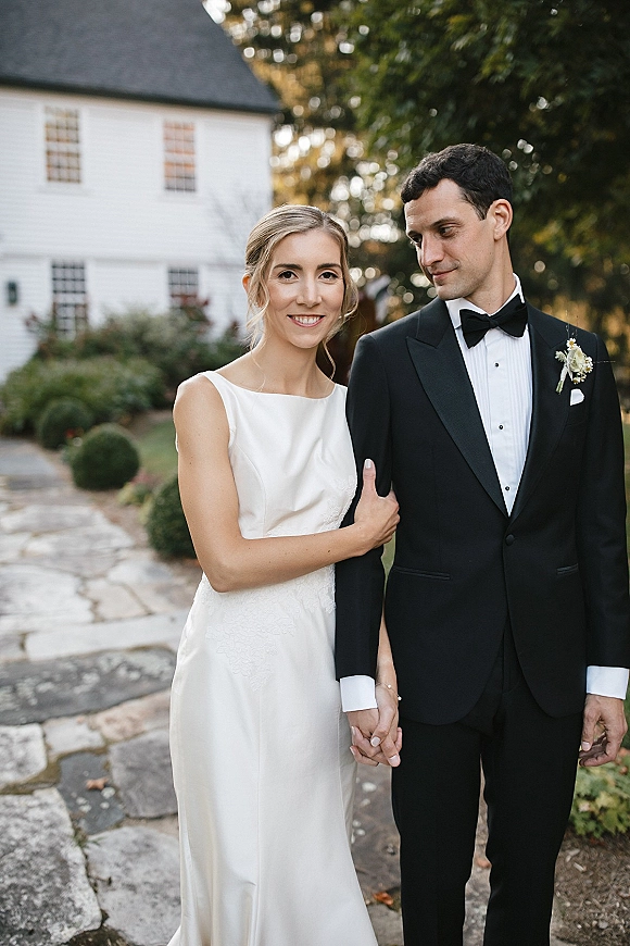 Couple portrait of bride and groom holding hands, bride in sleeveless wedding dress and groom in tuxedo on a stone path by a white house