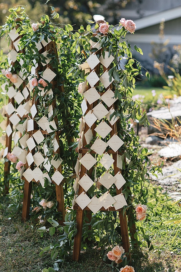 Wedding escort display with white calligraphy escort cards hanging on wooden lattice panels, greenery garland, and blush roses on a garden lawn