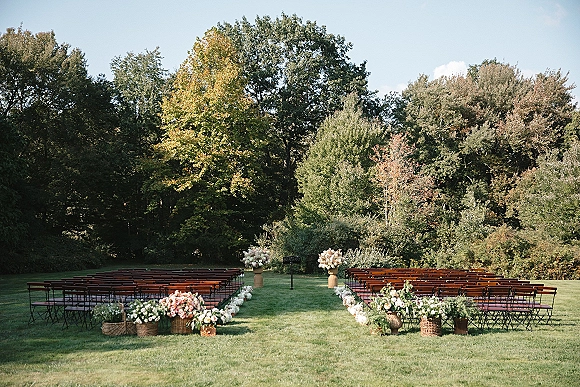 Outdoor ceremony setup with wood benches lining a center aisle, wicker baskets of flowers, and floral markers on a grassy lawn by trees