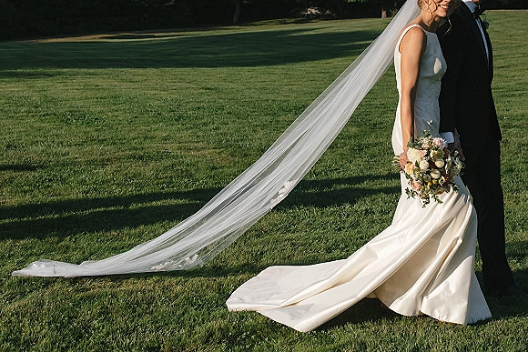 Couple portrait of bride and groom with long veil blowing behind her, bride holding bouquet on a sunlit park lawn with trees