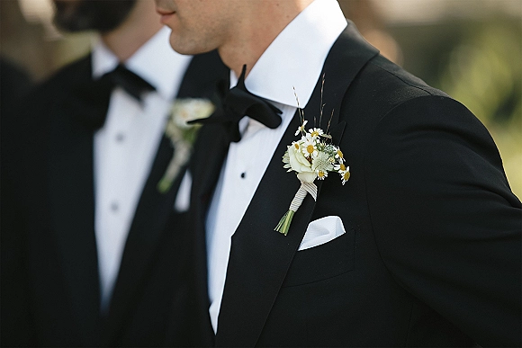 Groom boutonniere with a white rose boutonniere and daisy flowers pinned to a black tuxedo lapel, outdoor greenery and blurred groomsman behind