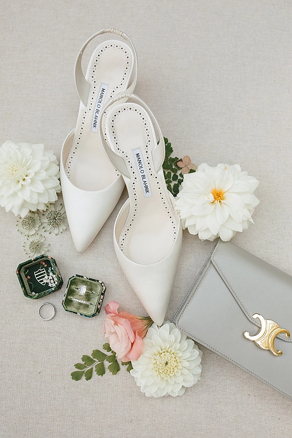 Bridal shoes styled with white slingback heels alongside ring boxes, engagement ring, and gray clutch on a linen backdrop