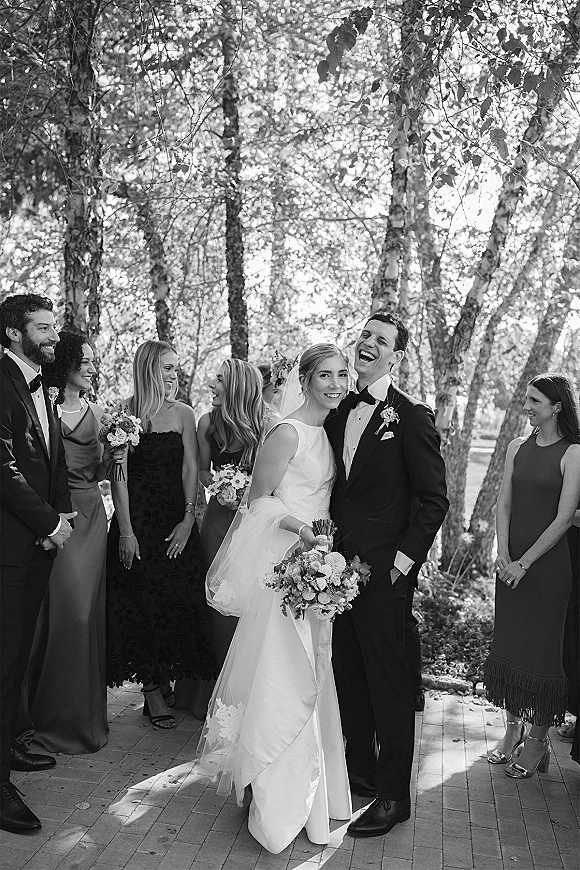 Wedding party photo of the bride and groom with bridesmaids, bride holding bouquet on a brick walkway in dappled garden light