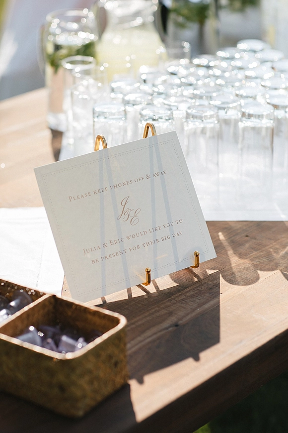 Wedding signage in a gold sign holder at a lemon water dispenser with glassware and ice bucket on a wood table in outdoor light