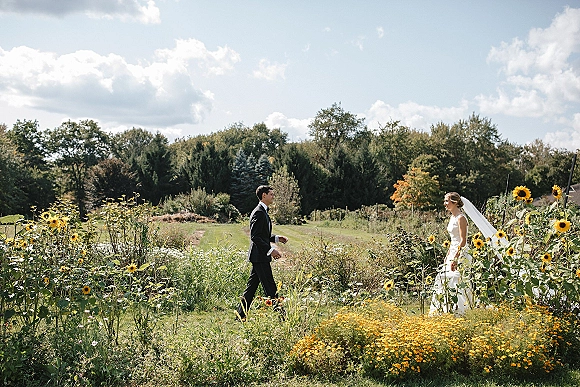 First look moment as bride in a white dress and veil approaches groom in black tuxedo on a meadow path under blue sky